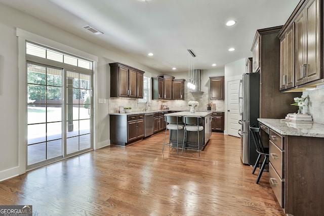 kitchen featuring wall chimney exhaust hood, a sink, stainless steel appliances, dark brown cabinetry, and a kitchen bar