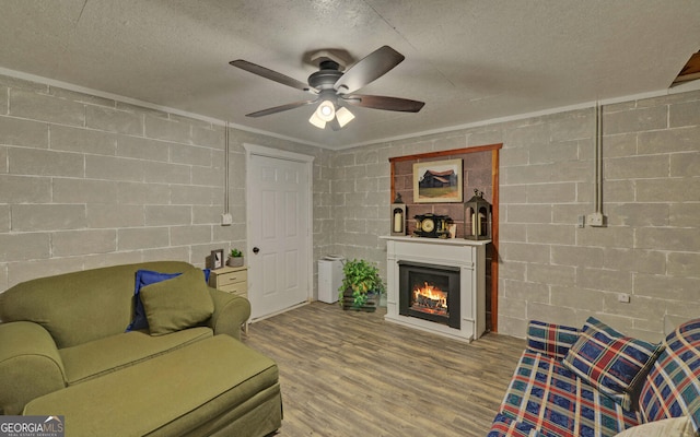 living area featuring a textured ceiling, ceiling fan, wood finished floors, a warm lit fireplace, and concrete block wall