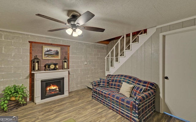 living area featuring a ceiling fan, wood finished floors, a textured ceiling, a warm lit fireplace, and concrete block wall