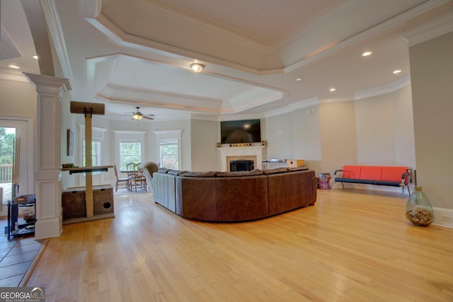 living area featuring light wood-style floors, ornate columns, a raised ceiling, a ceiling fan, and crown molding