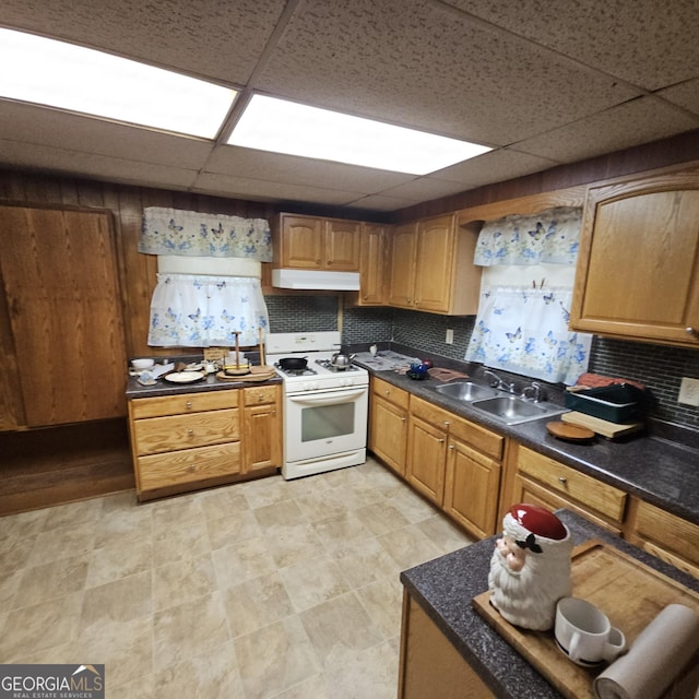 kitchen with a sink, gas range gas stove, under cabinet range hood, a drop ceiling, and dark countertops