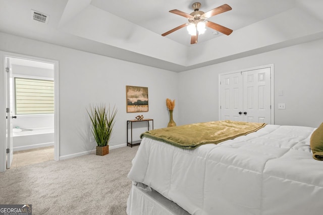 bedroom featuring light colored carpet, a tray ceiling, baseboards, a closet, and ensuite bath