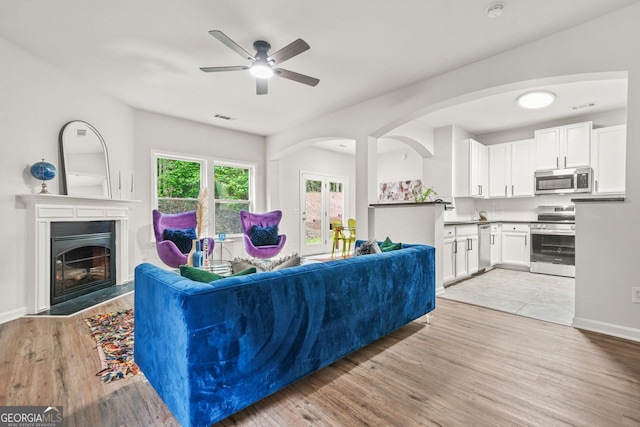 living area featuring a ceiling fan, a glass covered fireplace, light wood finished floors, arched walkways, and baseboards