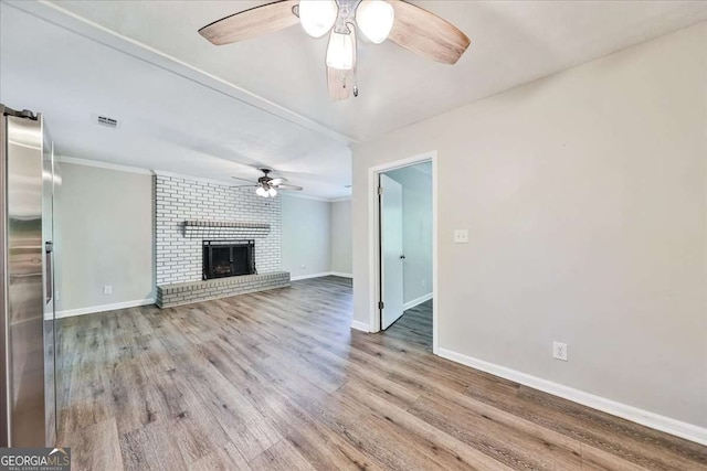 unfurnished living room with ceiling fan, baseboards, wood finished floors, crown molding, and a brick fireplace
