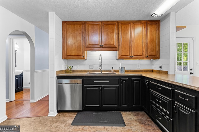kitchen featuring stainless steel dishwasher, a sink, arched walkways, brown cabinetry, and light countertops