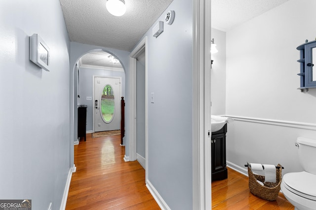 hall featuring arched walkways, a textured ceiling, light wood finished floors, baseboards, and ornamental molding