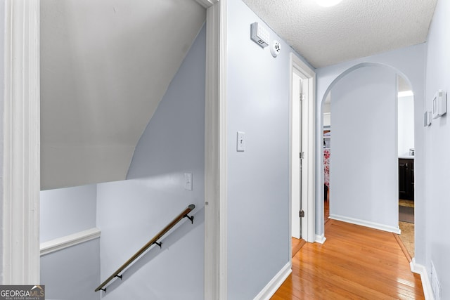 corridor with an upstairs landing, light wood-style floors, arched walkways, baseboards, and a textured ceiling