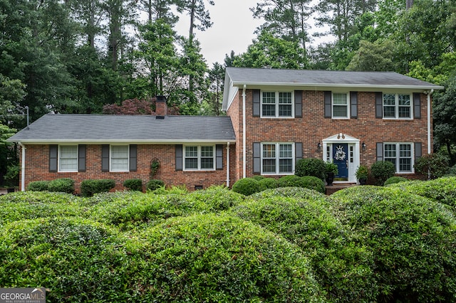 colonial inspired home featuring a chimney, brick siding, and view of wooded area