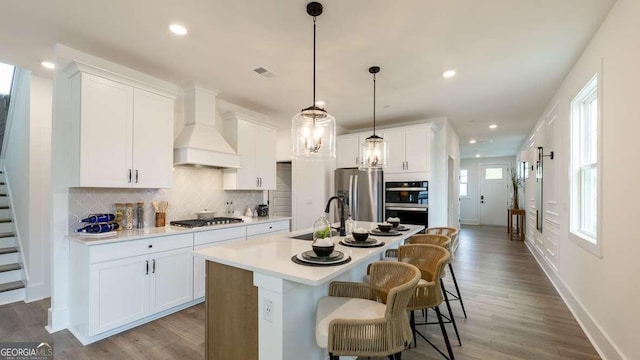 kitchen featuring a sink, appliances with stainless steel finishes, tasteful backsplash, an island with sink, and premium range hood