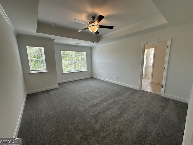 empty room with a tray ceiling, a ceiling fan, ornamental molding, dark carpet, and baseboards