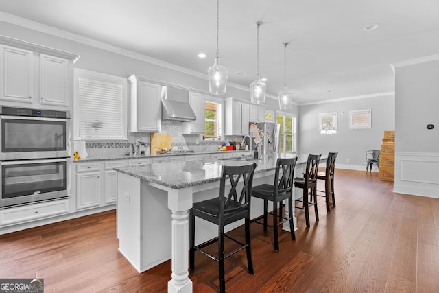 132 Shellbark Dr, Mcdonough, GA 30252 - kitchen featuring appliances with stainless steel finishes, a center island with sink, wall chimney range hood, white cabinetry, and ornamental molding