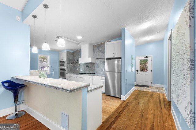 6567 Tahiti Way, Flowery Branch, GA 30542 - kitchen with stainless steel appliances, wall chimney range hood, tasteful backsplash, a peninsula, and light wood finished floors
