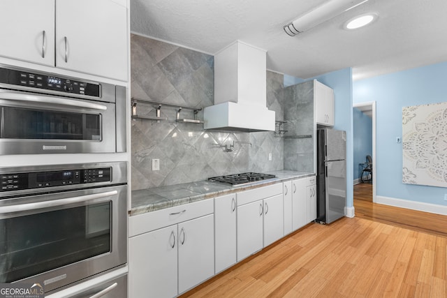 6567 Tahiti Way, Flowery Branch, GA 30542 - kitchen featuring stainless steel appliances, wall chimney exhaust hood, light wood-style flooring, backsplash, and white cabinetry