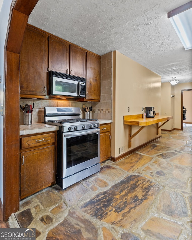 1290 Shadowlawn Dr NE, Conyers, GA 30012 - kitchen with appliances with stainless steel finishes, stone flooring, decorative backsplash, a textured ceiling, and brown cabinetry