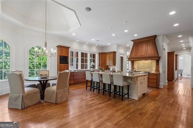 4634 Whitestone Way, Suwanee, GA 30024 - kitchen featuring custom exhaust hood, brown cabinets, a center island with sink, ornamental molding, and wood finished floors