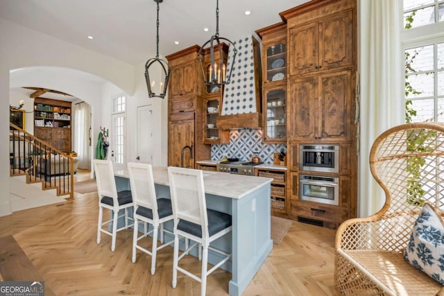 11277 Serenbe Ln, Chattahoochee Hills, GA 30268 - kitchen with stainless steel oven, arched walkways, brown cabinets, an island with sink, and decorative light fixtures