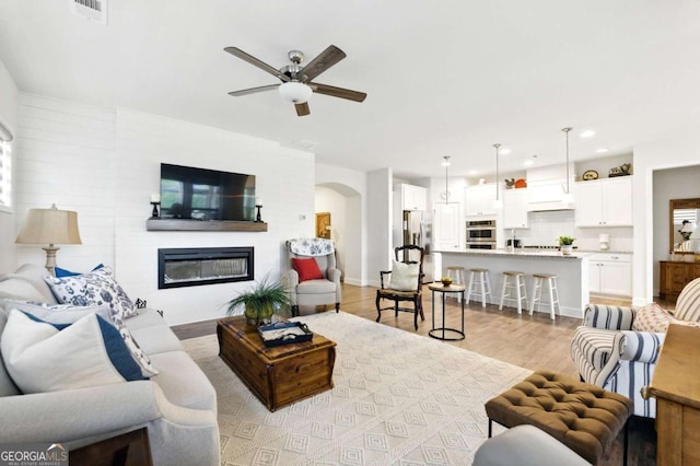 5744 Cypress Bluff Ln, Hoschton, GA 30548 - living room featuring arched walkways, a ceiling fan, a glass covered fireplace, light wood finished floors, and recessed lighting