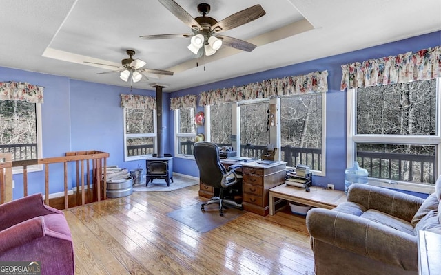 home office with ceiling fan, light hardwood / wood-style floors, a raised ceiling, and a wood stove