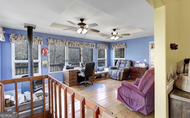 home office with ceiling fan, light wood-type flooring, a raised ceiling, and a wood stove