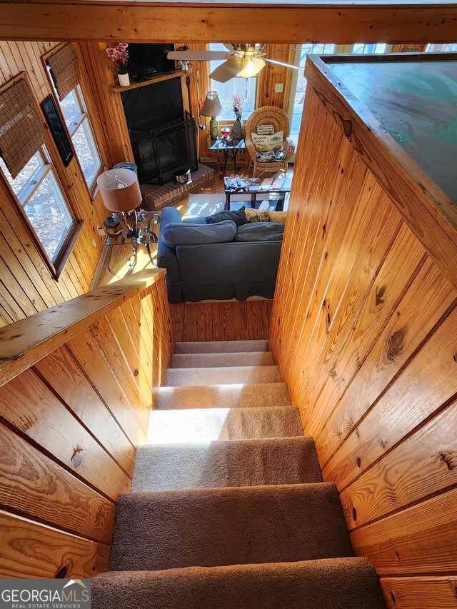 staircase with a fireplace, wood walls, ceiling fan, and a wealth of natural light