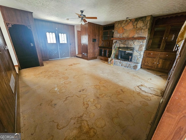 unfurnished living room featuring ceiling fan, a textured ceiling, wooden walls, and a stone fireplace