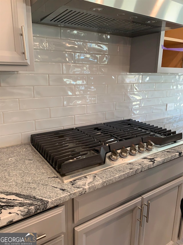 kitchen featuring stainless steel gas stovetop, backsplash, wall chimney range hood, gray cabinetry, and light stone countertops