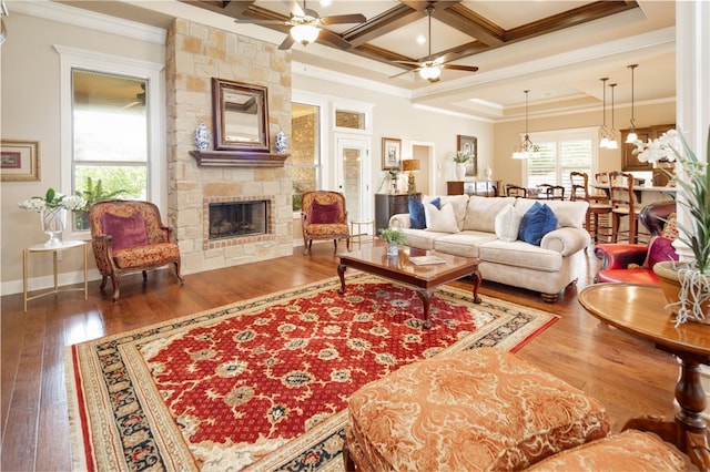 living room featuring dark hardwood / wood-style flooring, coffered ceiling, ceiling fan with notable chandelier, crown molding, and a stone fireplace