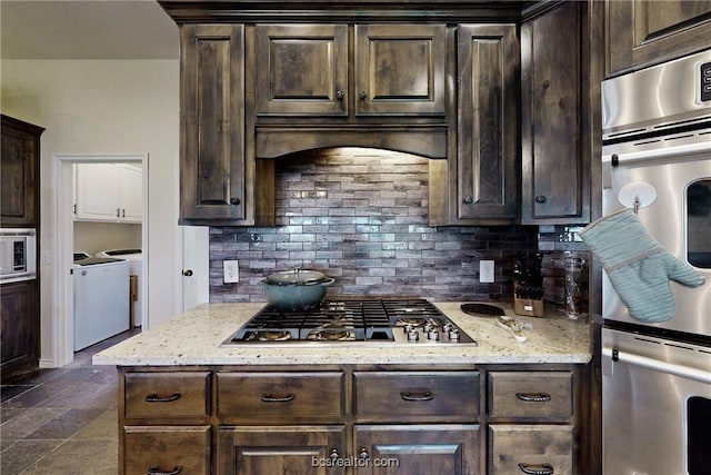 7576 River Ridge Drive, College Station, TX 77845 - kitchen with appliances with stainless steel finishes, washing machine and dryer, backsplash, dark brown cabinets, and light stone counters