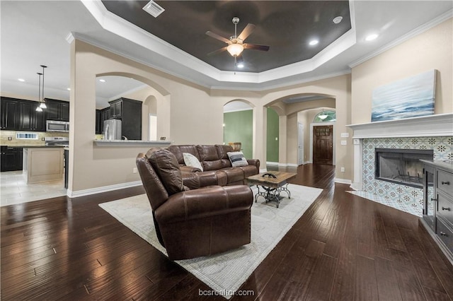 living room featuring dark wood-type flooring, a tile fireplace, a raised ceiling, crown molding, and ceiling fan