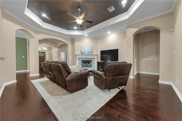 living room featuring dark wood-type flooring, a stone fireplace, a raised ceiling, crown molding, and ceiling fan