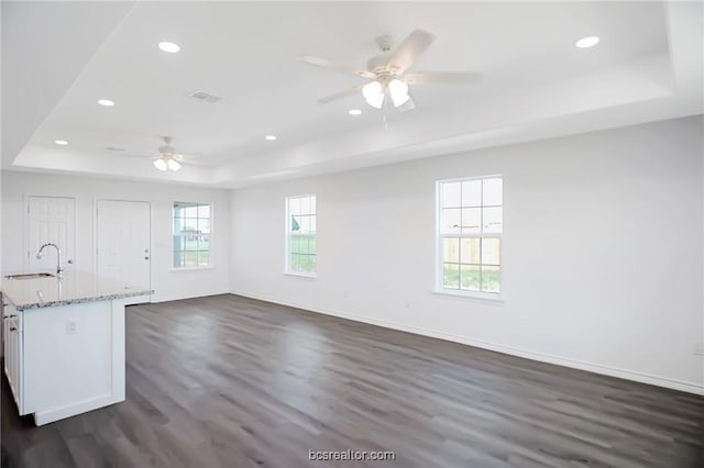 unfurnished living room featuring a raised ceiling, ceiling fan, dark wood-type flooring, and a healthy amount of sunlight