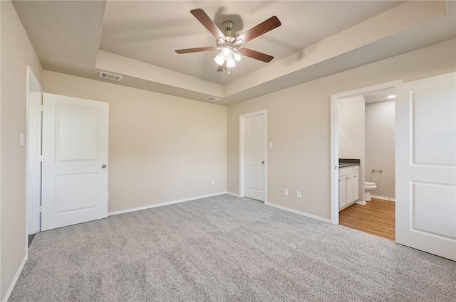 unfurnished bedroom featuring a raised ceiling, ceiling fan, light colored carpet, and ensuite bath