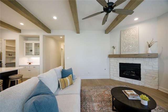 living room featuring beam ceiling, light hardwood / wood-style floors, a stone fireplace, and ceiling fan