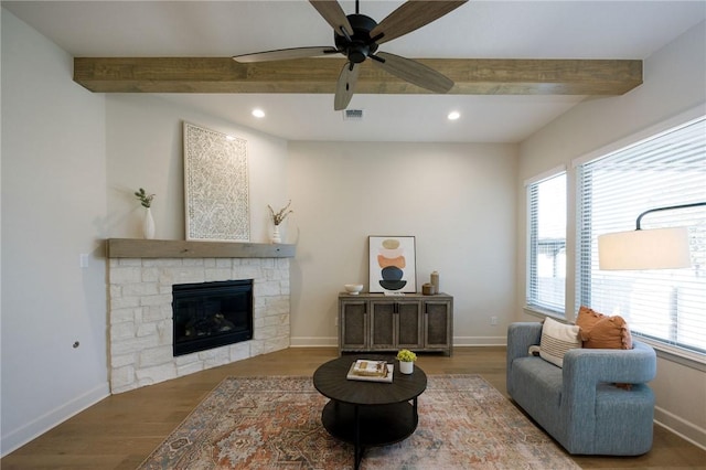 living room featuring beamed ceiling, a stone fireplace, dark wood-type flooring, and ceiling fan