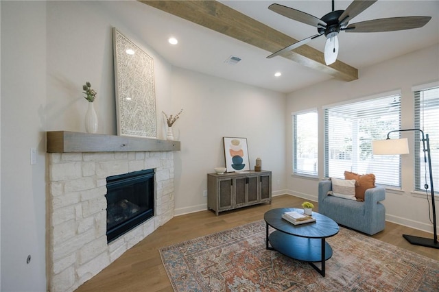 living area featuring hardwood / wood-style floors, ceiling fan, beam ceiling, and a stone fireplace