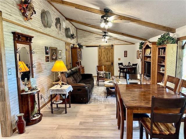 dining area with ceiling fan, a barn door, light wood-style floors, wood walls, and vaulted ceiling with beams