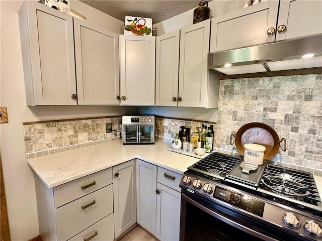 kitchen with under cabinet range hood, backsplash, light stone countertops, and gas range
