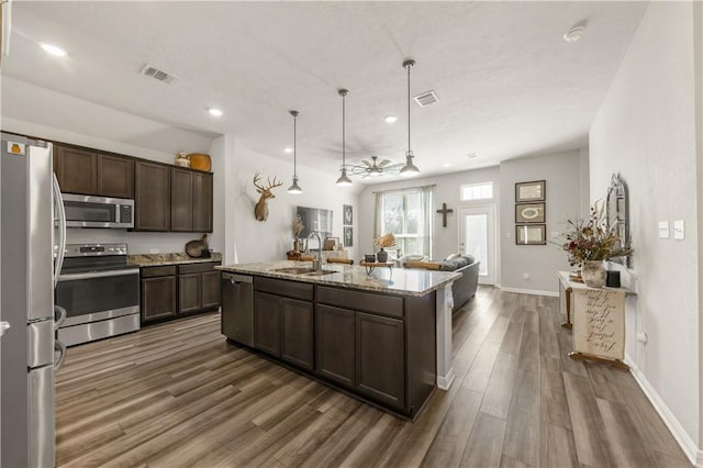 kitchen featuring an island with sink, appliances with stainless steel finishes, light stone countertops, pendant lighting, and a sink