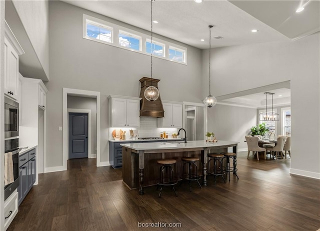 kitchen with dark wood-type flooring, white cabinets, a high ceiling, hanging light fixtures, and an island with sink