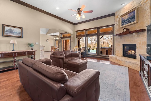 living area with ceiling fan, arched walkways, a stone fireplace, wood finished floors, and crown molding