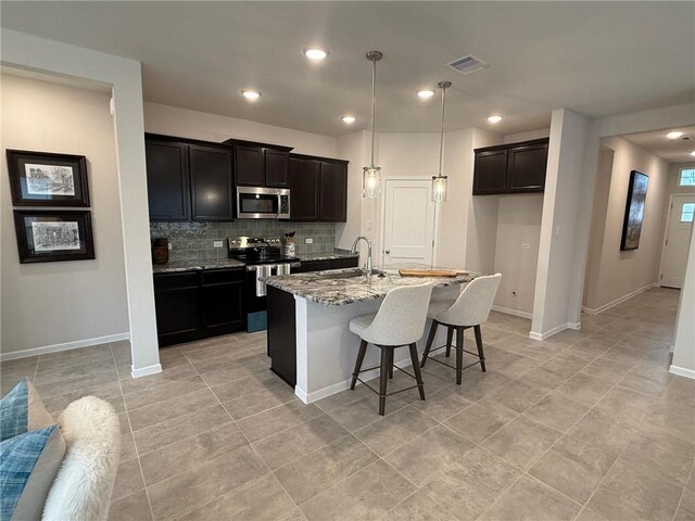 kitchen featuring dark stone counters, a center island with sink, appliances with stainless steel finishes, tasteful backsplash, and decorative light fixtures