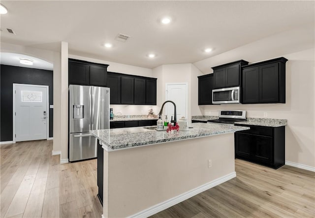 kitchen with sink, light hardwood / wood-style flooring, an island with sink, appliances with stainless steel finishes, and light stone counters