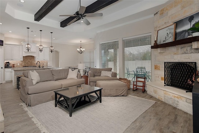 living room with crown molding, ceiling fan with notable chandelier, light hardwood / wood-style floors, a stone fireplace, and beamed ceiling