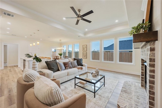 living room with a brick fireplace, ceiling fan with notable chandelier, a raised ceiling, and light wood-type flooring