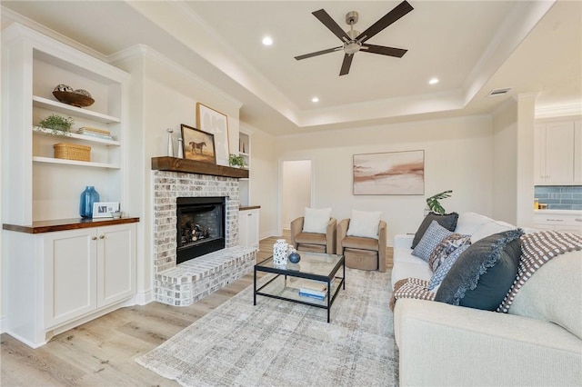 living room featuring crown molding, ceiling fan, a raised ceiling, light wood-type flooring, and a brick fireplace