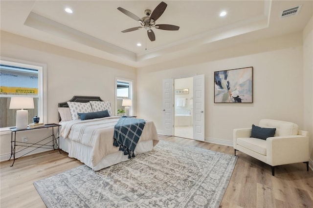 bedroom featuring ceiling fan, light hardwood / wood-style flooring, ensuite bathroom, and a raised ceiling