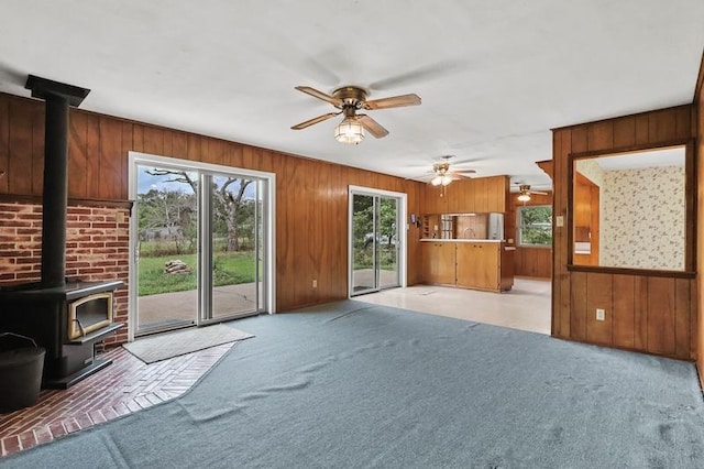 unfurnished living room featuring light carpet, a wood stove, wood walls, and a ceiling fan