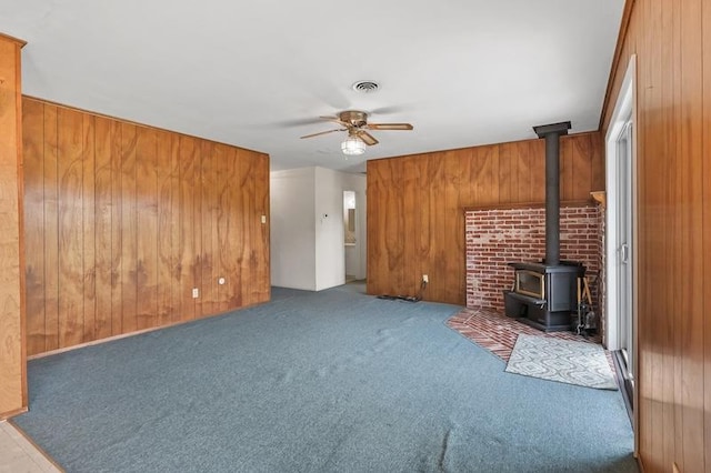 unfurnished living room featuring wood walls, a wood stove, visible vents, carpet floors, and a ceiling fan