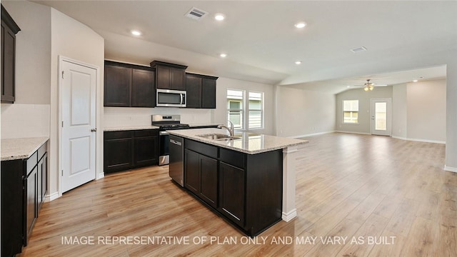 kitchen with stainless steel appliances, an island with sink, sink, light stone counters, and light hardwood / wood-style flooring