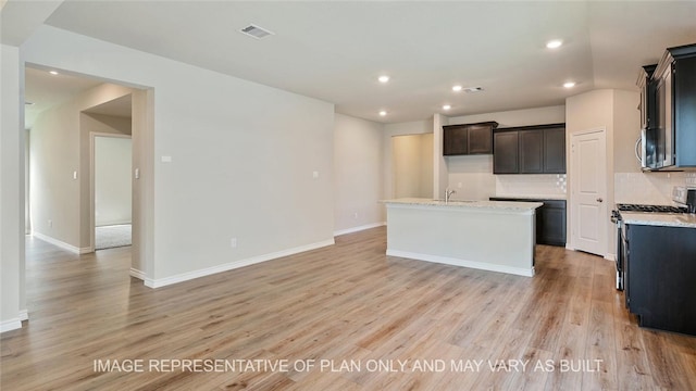 kitchen featuring a center island with sink, appliances with stainless steel finishes, decorative backsplash, light hardwood / wood-style flooring, and light stone counters